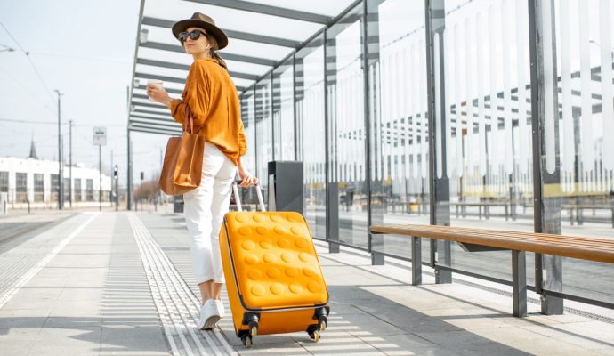 Young female traveler with a luggage at the transport stop Young female traveler walking with a yellow suitcase at the modern transport stop outdoors, back view. Concept of an urban transportation and travel